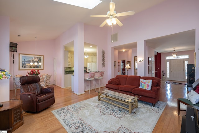 living room featuring light wood finished floors, a skylight, visible vents, high vaulted ceiling, and ceiling fan with notable chandelier