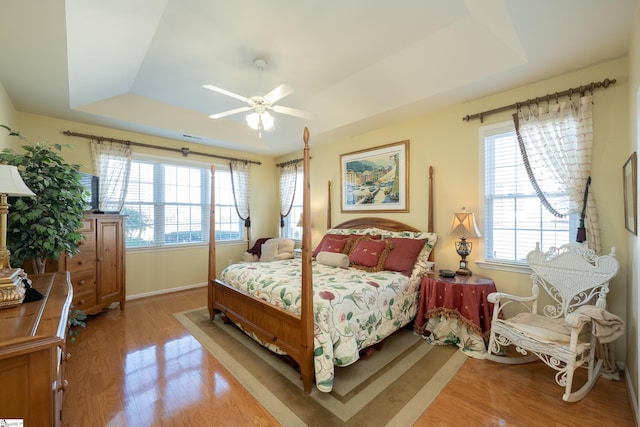 bedroom with light wood-style floors, a tray ceiling, multiple windows, and a ceiling fan