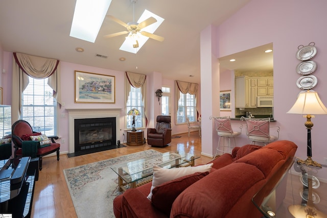 living room with a skylight, visible vents, a fireplace with flush hearth, light wood-type flooring, and high vaulted ceiling