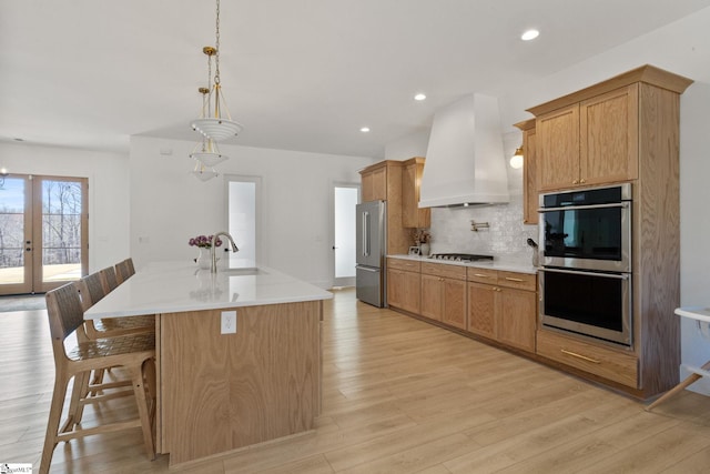 kitchen with stainless steel appliances, a sink, light wood-style floors, decorative backsplash, and wall chimney exhaust hood