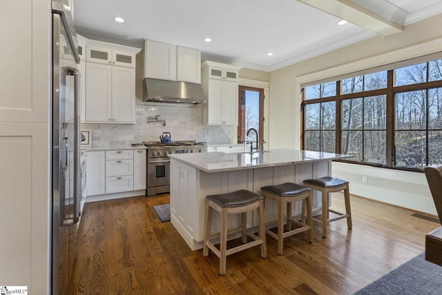 kitchen featuring white cabinets, light stone countertops, high end stove, under cabinet range hood, and backsplash