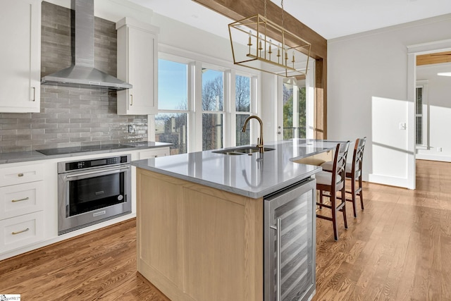 kitchen featuring beverage cooler, black electric cooktop, wall chimney range hood, stainless steel oven, and a sink