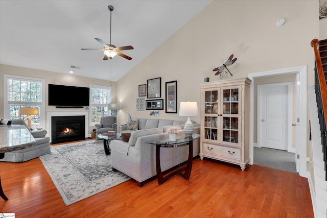 living area featuring ceiling fan, high vaulted ceiling, visible vents, a lit fireplace, and light wood finished floors