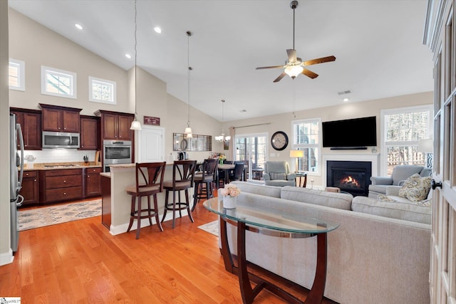 living area featuring ceiling fan, high vaulted ceiling, visible vents, light wood finished floors, and a glass covered fireplace