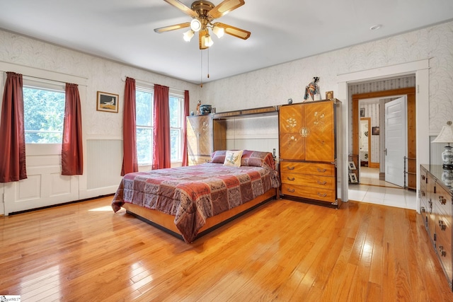 bedroom featuring a wainscoted wall, light wood-type flooring, a ceiling fan, and wallpapered walls