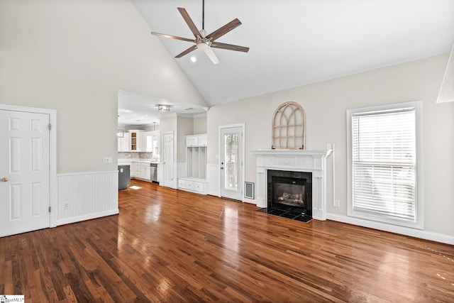 unfurnished living room featuring a tiled fireplace, wainscoting, ceiling fan, dark wood-type flooring, and high vaulted ceiling