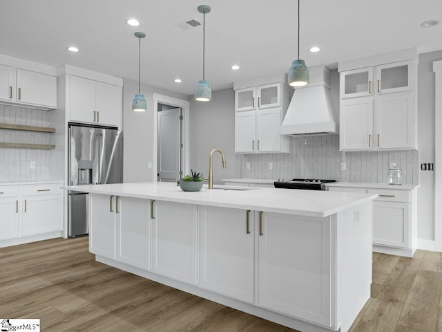 kitchen with stainless steel refrigerator with ice dispenser, light wood finished floors, visible vents, a sink, and premium range hood