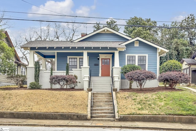 bungalow with covered porch, brick siding, stairs, roof with shingles, and a chimney