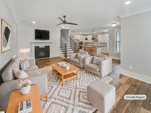 living area with a glass covered fireplace, stairway, wood finished floors, crown molding, and recessed lighting