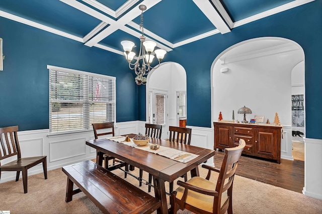 carpeted dining space with arched walkways, a wainscoted wall, coffered ceiling, beam ceiling, and an inviting chandelier