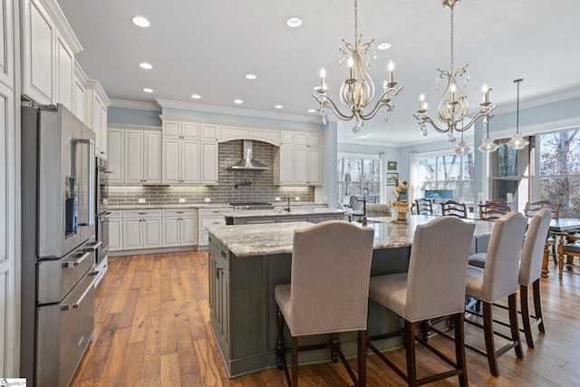 kitchen featuring a spacious island, wall chimney exhaust hood, wood-type flooring, appliances with stainless steel finishes, and ornamental molding