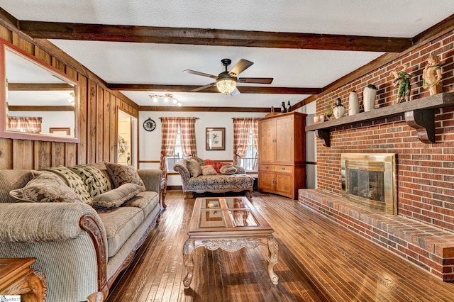 living area with a ceiling fan, hardwood / wood-style flooring, a textured ceiling, a brick fireplace, and beam ceiling