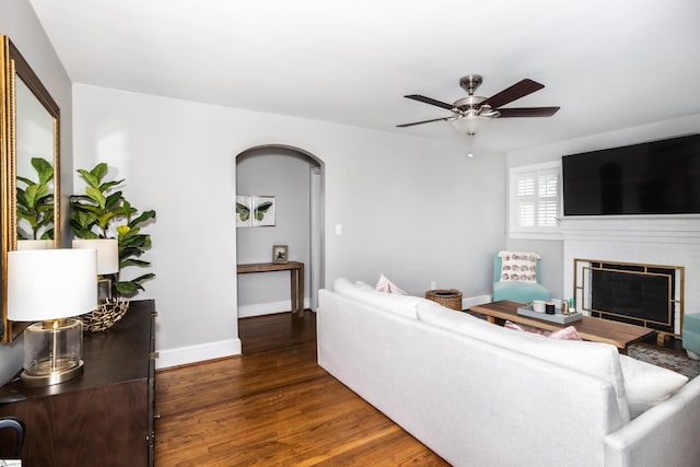 living room with arched walkways, ceiling fan, wood finished floors, baseboards, and a brick fireplace