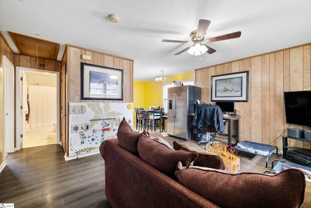 living room featuring ceiling fan with notable chandelier, dark wood-type flooring, wood walls, and attic access