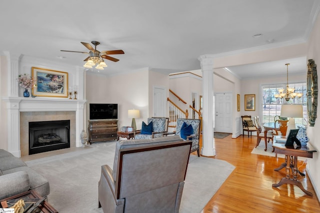 living room featuring ceiling fan with notable chandelier, stairway, light wood finished floors, a tiled fireplace, and ornate columns