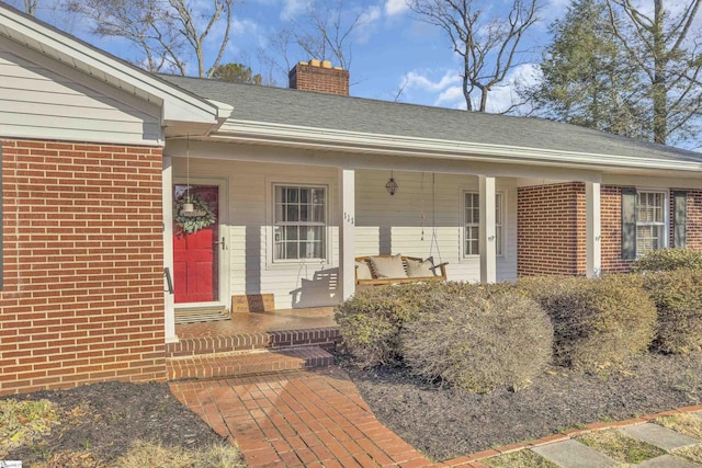 doorway to property featuring covered porch, roof with shingles, brick siding, and a chimney