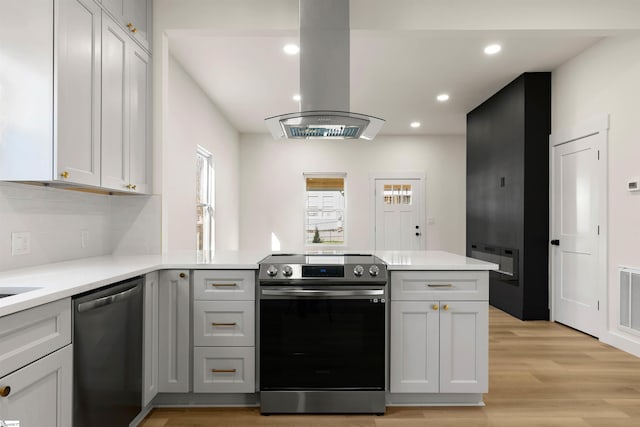 kitchen with appliances with stainless steel finishes, a peninsula, island exhaust hood, light countertops, and light wood-type flooring