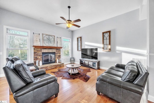 living area with ceiling fan, a stone fireplace, wood-type flooring, and baseboards