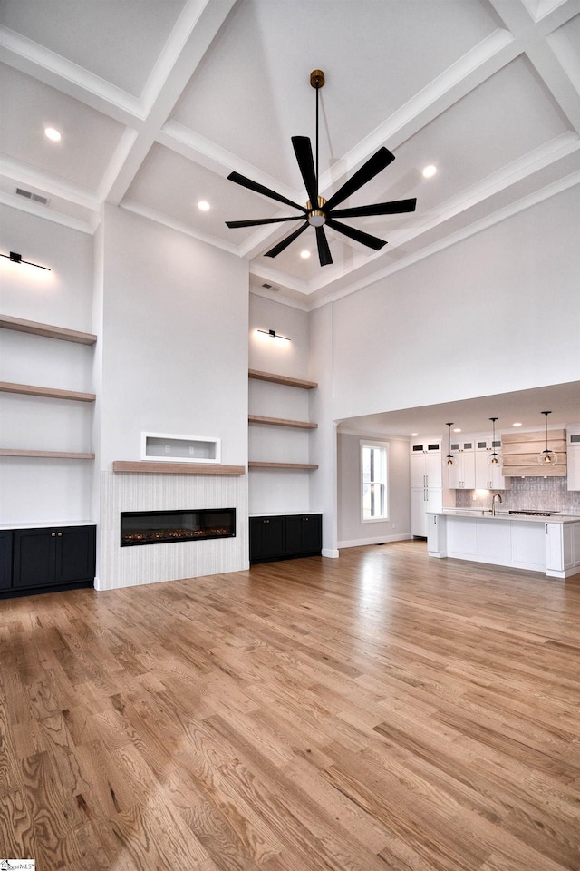 unfurnished living room with coffered ceiling, a ceiling fan, a glass covered fireplace, light wood-type flooring, and beam ceiling