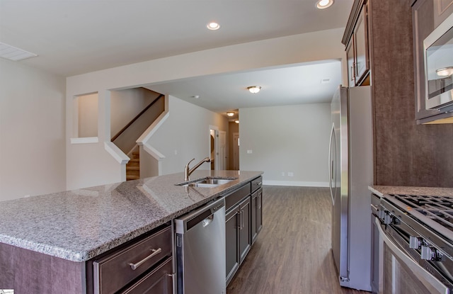 kitchen featuring a kitchen island with sink, dark wood-type flooring, a sink, appliances with stainless steel finishes, and light stone countertops