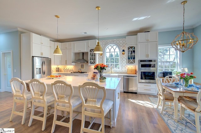 kitchen with stainless steel appliances, white cabinetry, light wood-style floors, wall chimney exhaust hood, and crown molding