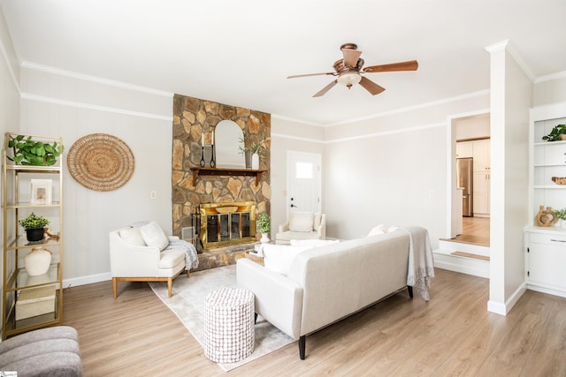 living area with a ceiling fan, baseboards, a stone fireplace, light wood-style floors, and crown molding