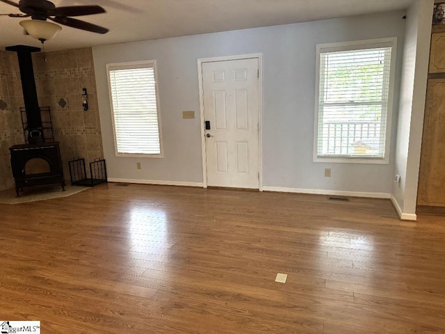 unfurnished living room featuring visible vents, baseboards, a wood stove, wood finished floors, and a ceiling fan