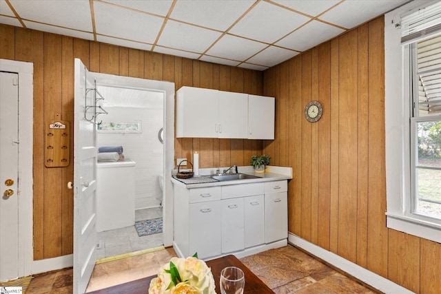 kitchen featuring a sink, a drop ceiling, white cabinetry, wooden walls, and light countertops