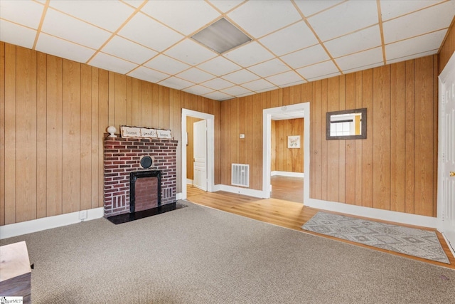 unfurnished living room featuring visible vents, a drop ceiling, wood walls, carpet flooring, and a brick fireplace