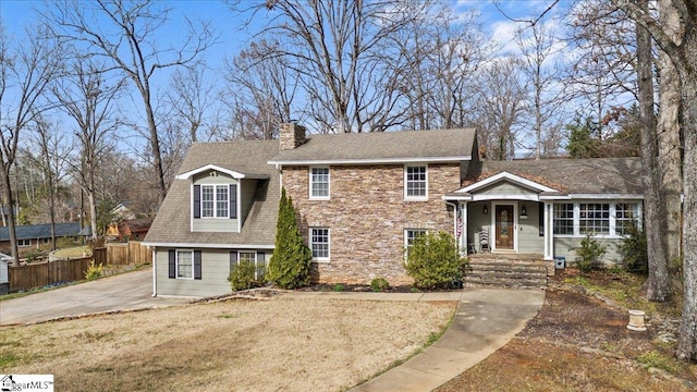 split level home with a front yard, fence, roof with shingles, a chimney, and stone siding