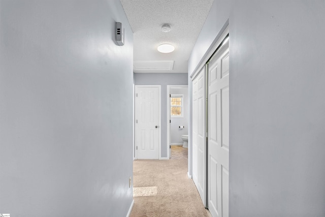 hall with baseboards, light colored carpet, attic access, and a textured ceiling