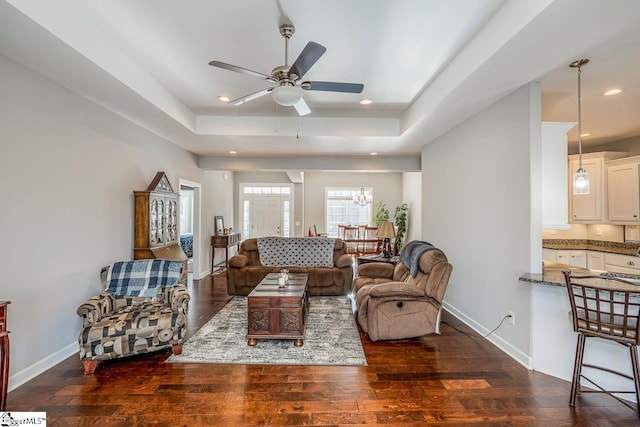 living room with a raised ceiling, ceiling fan with notable chandelier, recessed lighting, baseboards, and dark wood-style flooring