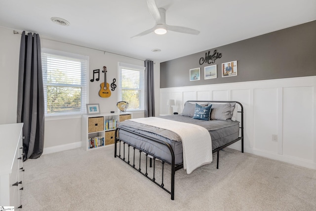 carpeted bedroom with visible vents, a decorative wall, ceiling fan, and a wainscoted wall