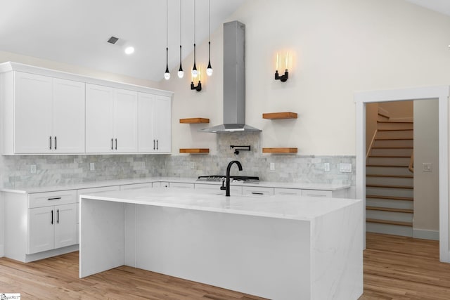 kitchen featuring light wood finished floors, open shelves, a kitchen island with sink, wall chimney range hood, and tasteful backsplash