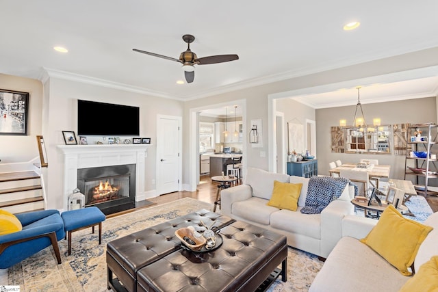 living area with stairway, light wood-type flooring, ornamental molding, ceiling fan with notable chandelier, and a glass covered fireplace