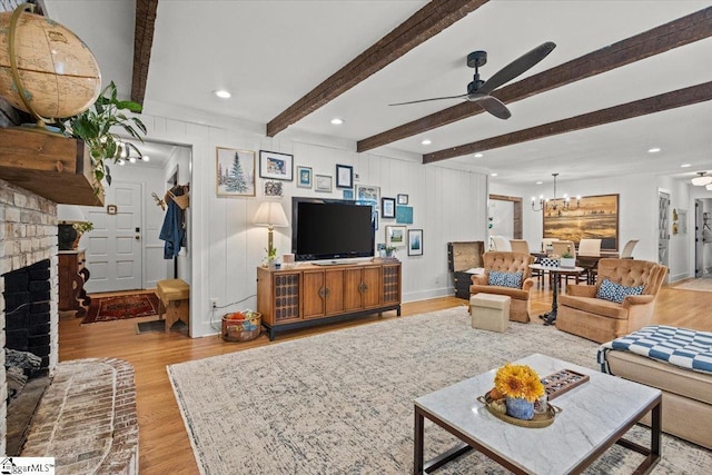 living area with beamed ceiling, recessed lighting, light wood-style floors, a brick fireplace, and ceiling fan with notable chandelier