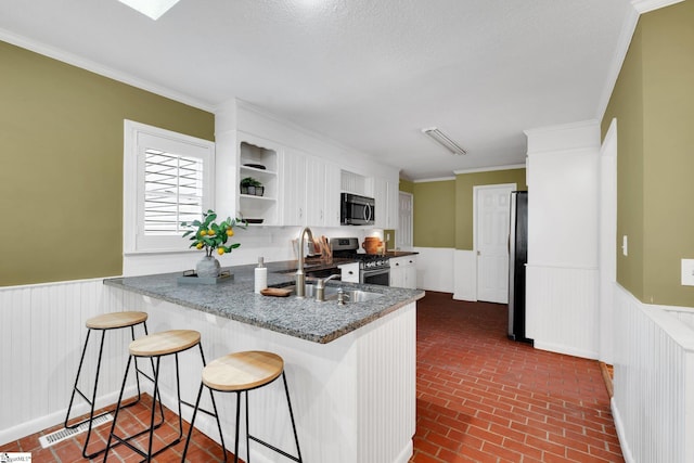 kitchen featuring white cabinetry, stainless steel appliances, a peninsula, wainscoting, and brick floor