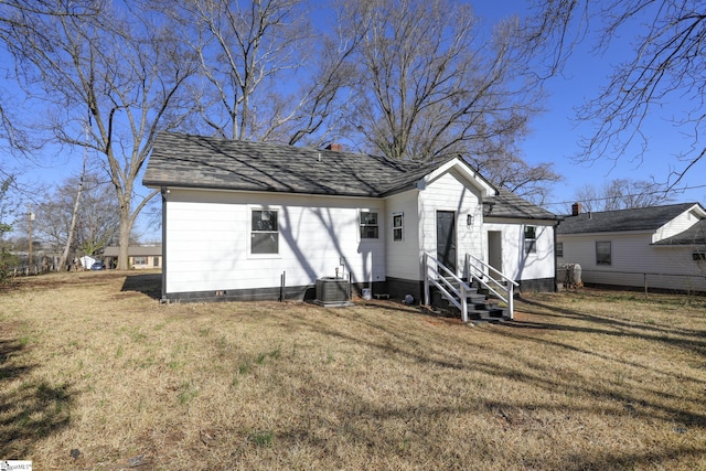 rear view of house featuring central air condition unit, entry steps, roof with shingles, a lawn, and a chimney