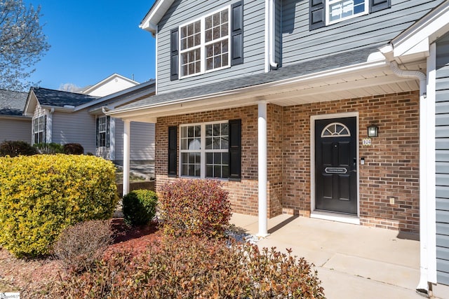 doorway to property featuring brick siding, a porch, and a shingled roof