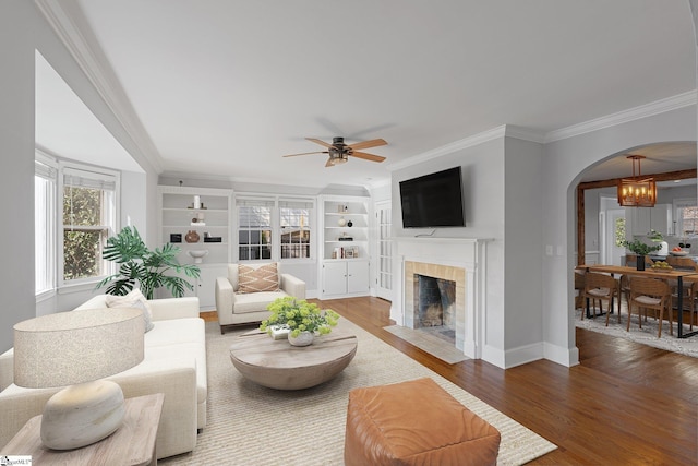 living area with ceiling fan with notable chandelier, wood finished floors, arched walkways, crown molding, and a tile fireplace