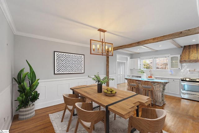 dining room with beam ceiling, ornamental molding, light wood-style floors, an inviting chandelier, and wainscoting