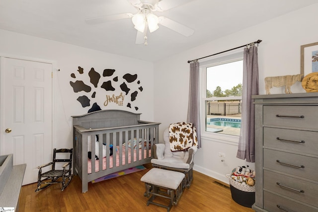 bedroom featuring visible vents, a crib, wood finished floors, baseboards, and ceiling fan