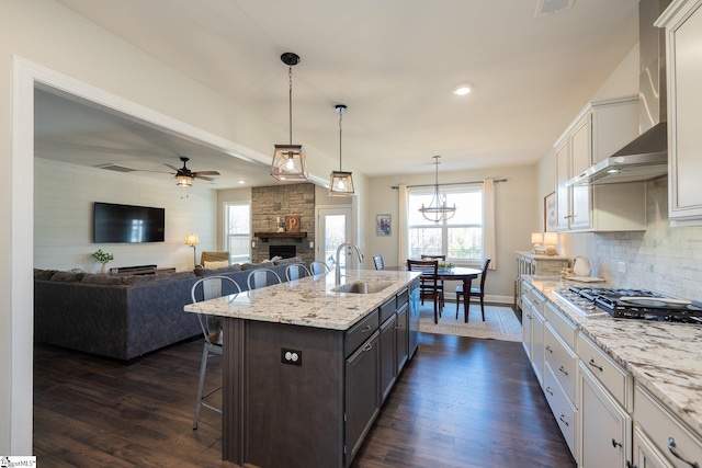 kitchen featuring wall chimney range hood, dark wood finished floors, a stone fireplace, a kitchen breakfast bar, and a sink