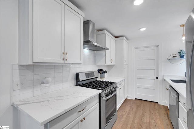 kitchen featuring wall chimney range hood, light wood-type flooring, decorative backsplash, stainless steel appliances, and white cabinetry