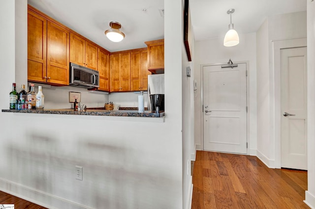 kitchen featuring wood finished floors, dark stone counters, hanging light fixtures, appliances with stainless steel finishes, and brown cabinets