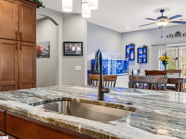 kitchen with light stone counters, a ceiling fan, arched walkways, a sink, and brown cabinets