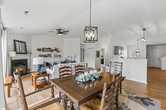 dining room featuring visible vents, ceiling fan with notable chandelier, a warm lit fireplace, wood finished floors, and vaulted ceiling