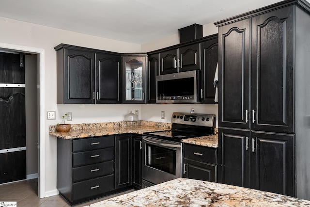 kitchen with light stone counters, appliances with stainless steel finishes, light tile patterned flooring, glass insert cabinets, and dark cabinets