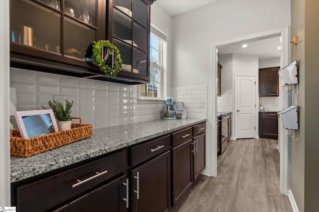 kitchen featuring tasteful backsplash, glass insert cabinets, light stone countertops, dark brown cabinetry, and light wood-style flooring