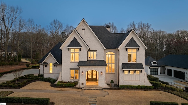 french country inspired facade featuring driveway, a standing seam roof, a chimney, a shingled roof, and brick siding